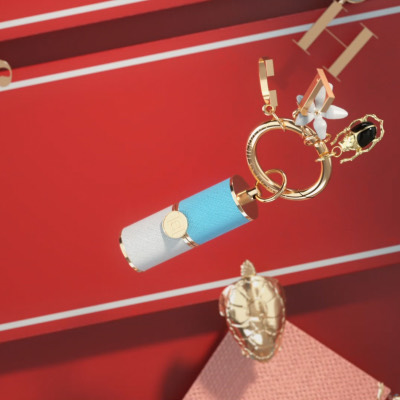 A blue and white lipstick case hanging from a gold ring charm against a red wall, a gold compact below it.
